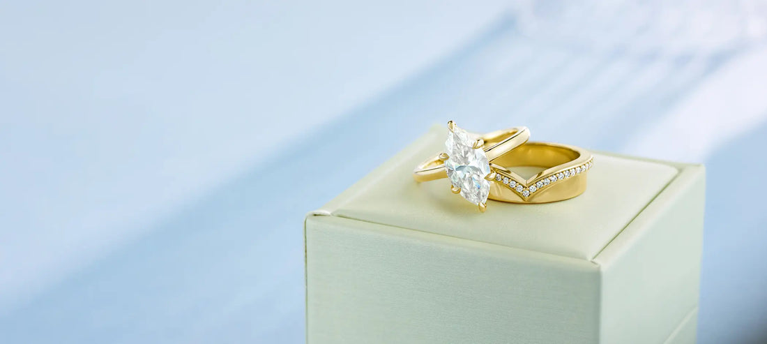 a gold marquise engagement ring and thick gold band on a pedestal against a blue background