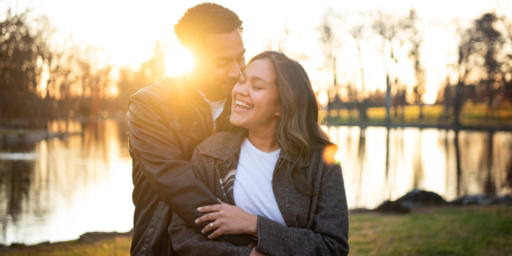 Couple embracing by a lake at sunset. Brand new love with oval solitaire diamond in a yellow gold setting.