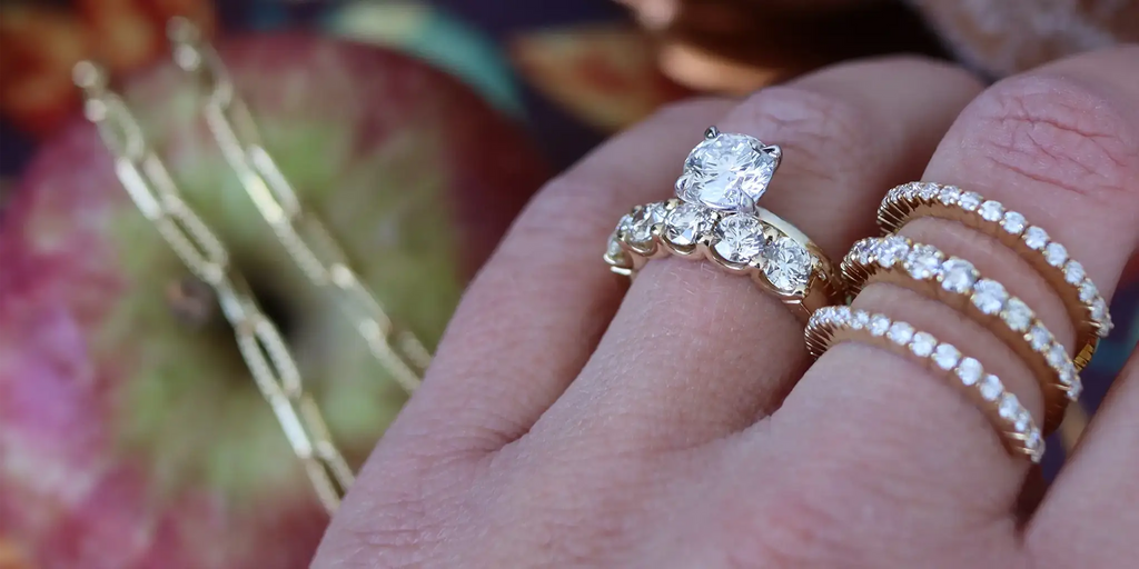 Engagement ring and wedding bands on woman's fingers. Apple and necklace in background.
